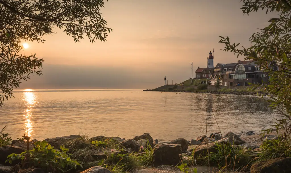 De vuurtoren van Urk in de avond met een ondergaande zon, op een rustige zomerdag, gezien vanaf het strand vanachter de boompjes.